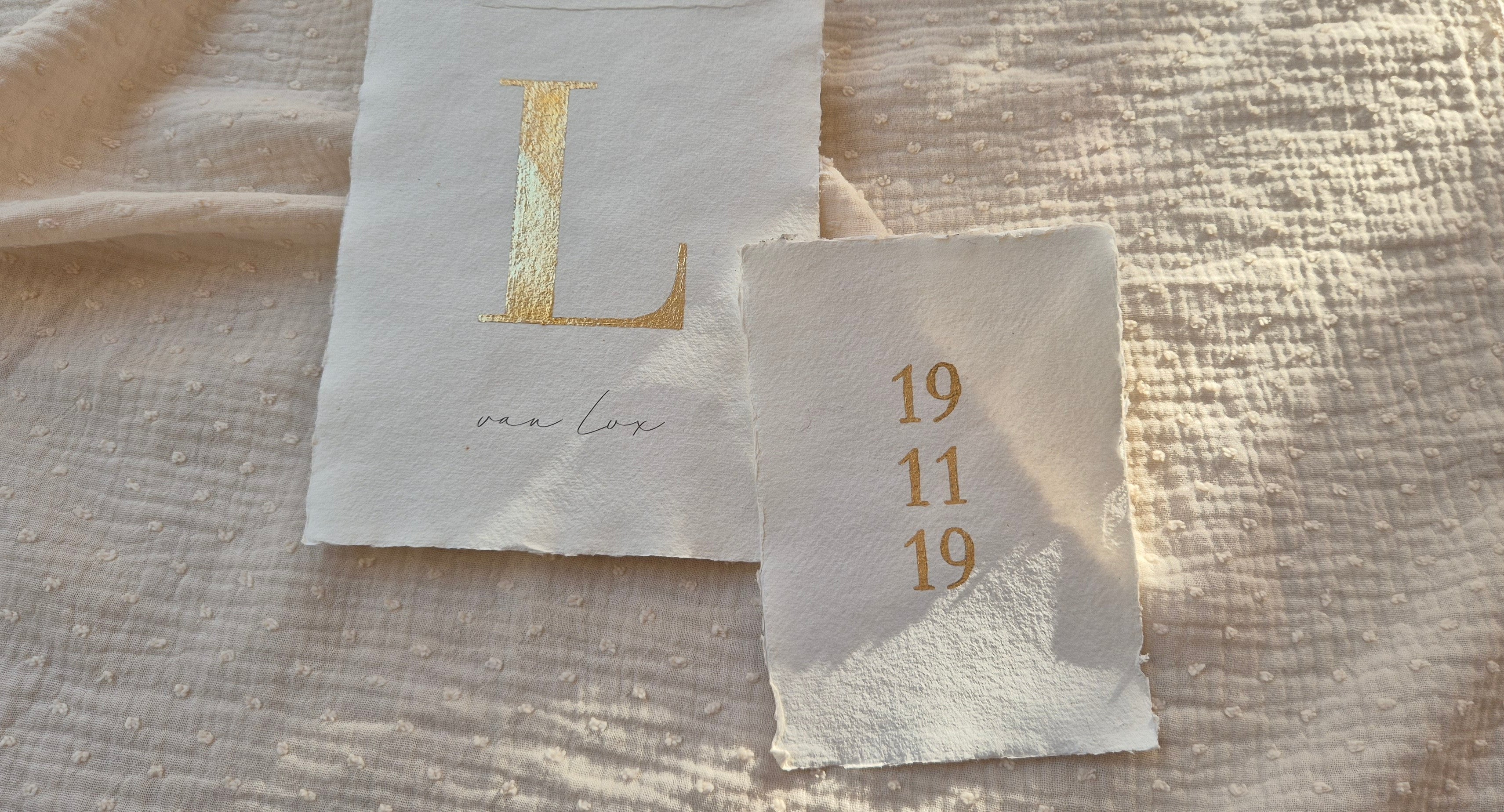 Handgemaakte letter- en datumposter op geschept papier met ruwe randen. Links een goudfolie letter “L” met daaronder een naam in fijne handgeschreven typografie, rechts een bijpassende poster met een geboortedatum in goudfolie. Gefotografeerd op een zachte, lichtgekleurde stoffen ondergrond met warm natuurlijk licht voor een rustige, ambachtelijke uitstraling.