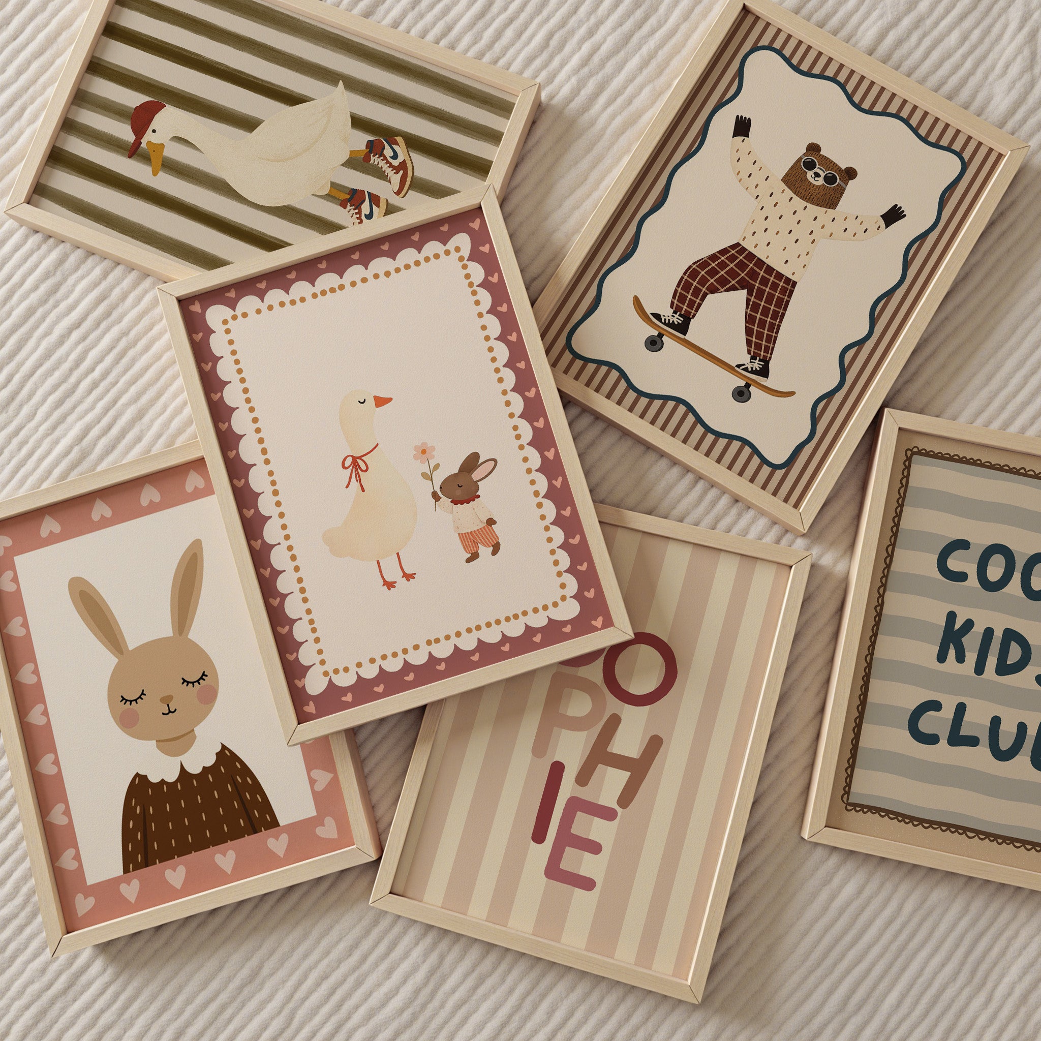 Zes houten fotolijsten liggen speels verspreid op een licht ribstof kleed. De illustraties hebben een zachte, retro kinderkamerstijl met warme beige, roze en bruine tinten: een gans met sneakers, een beer op een skateboard, een konijnportret, een gans met een konijntje, een typografische poster met ‘SOPHIE’ en een poster met de tekst ‘COOL KIDS CLUB’