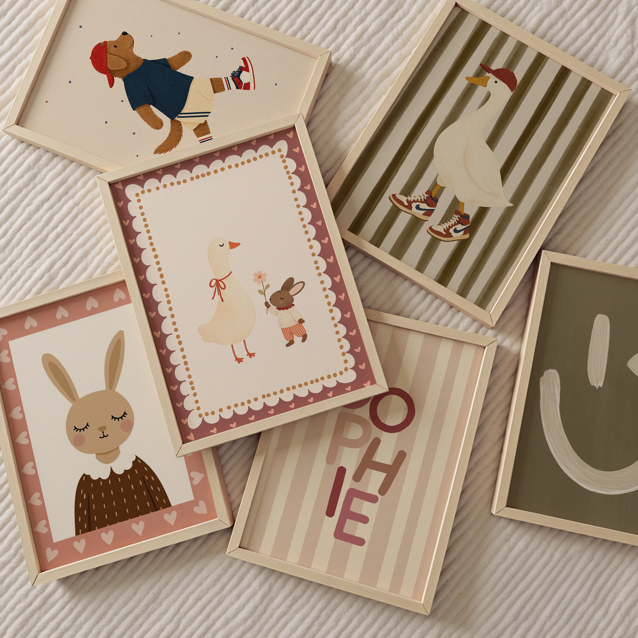 Zes houten fotolijsten liggen speels verspreid op een licht ribstof kleed. De illustraties hebben een zachte, retro kinderkamerstijl met warme beige, roze en bruine tinten: een gans met sneakers, een stoere hond, een konijnportret, een gans met een konijntje, een typografische poster met ‘SOPHIE’ en een poster met smiley