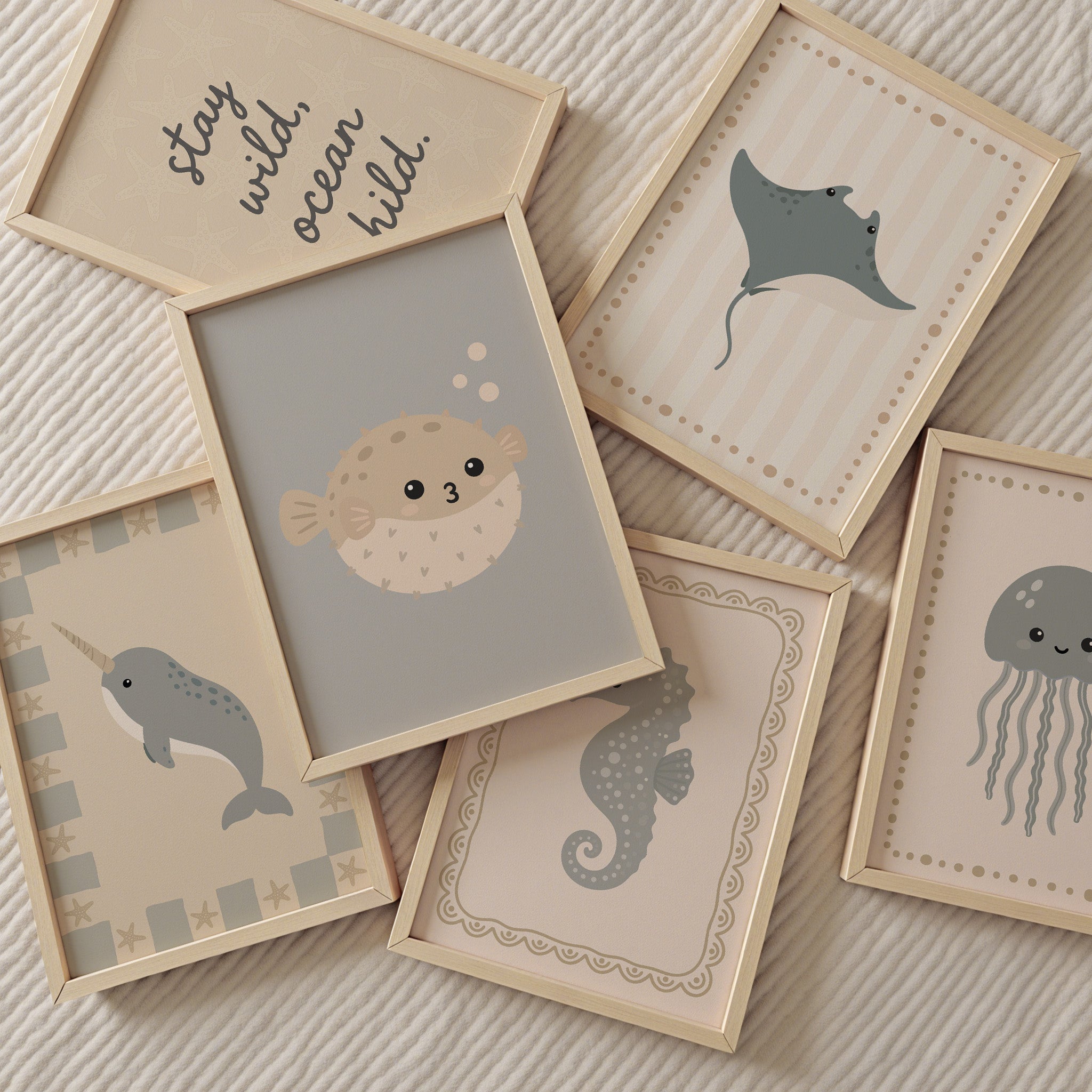 Zes ingelijste posters met een zacht zeethema liggen verspreid op een beige ribstof. De posters tonen schattige illustraties van een kogelvis, pijlstaartrog, narwal, zeepaardje en kwal in neutrale pastelkleuren. Op één poster staat de tekst: “stay wild ocean child”. De lichte houten lijsten en rustige kleuren geven een warme, speelse uitstraling die geschikt is voor een baby- of kinderkamer.